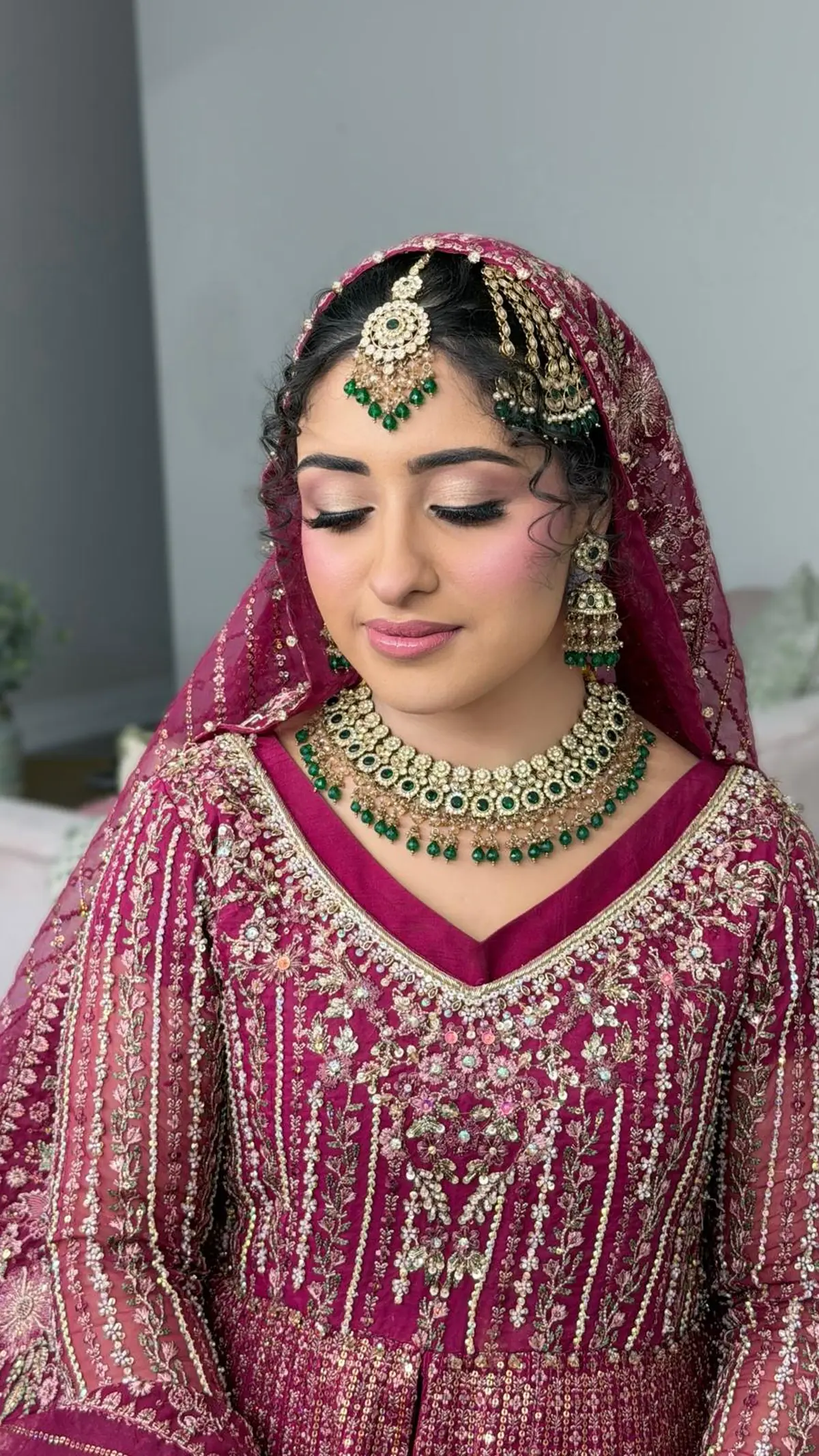 Bride in fuchsia with an emerald kundan necklace, jhoomar and maang tikka, soft curls framing her face under an embroidered dupatta.