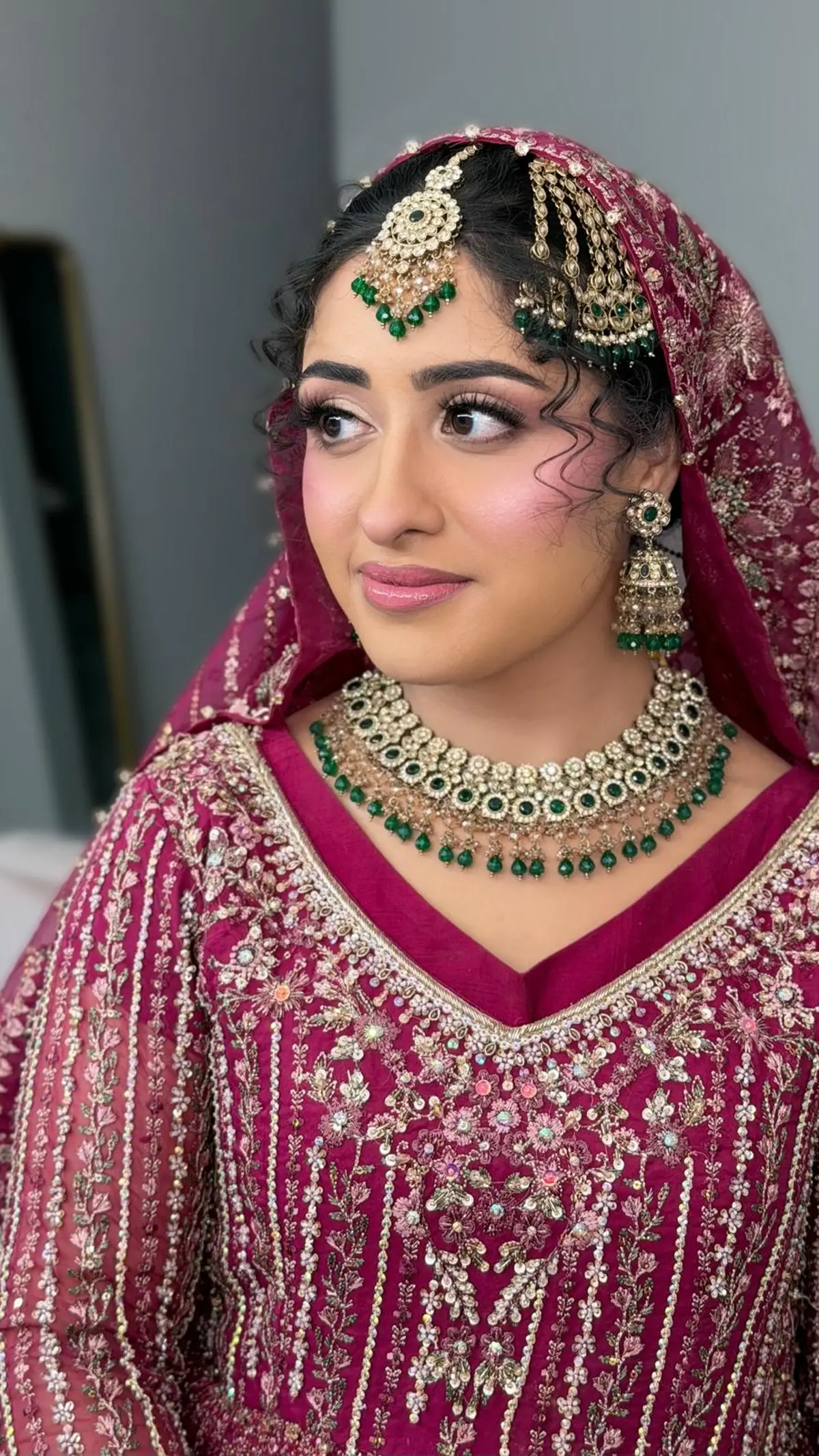 The fuchsia bride glancing aside, emerald jhumkas and soft pink lips, curls loose under her dupatta.