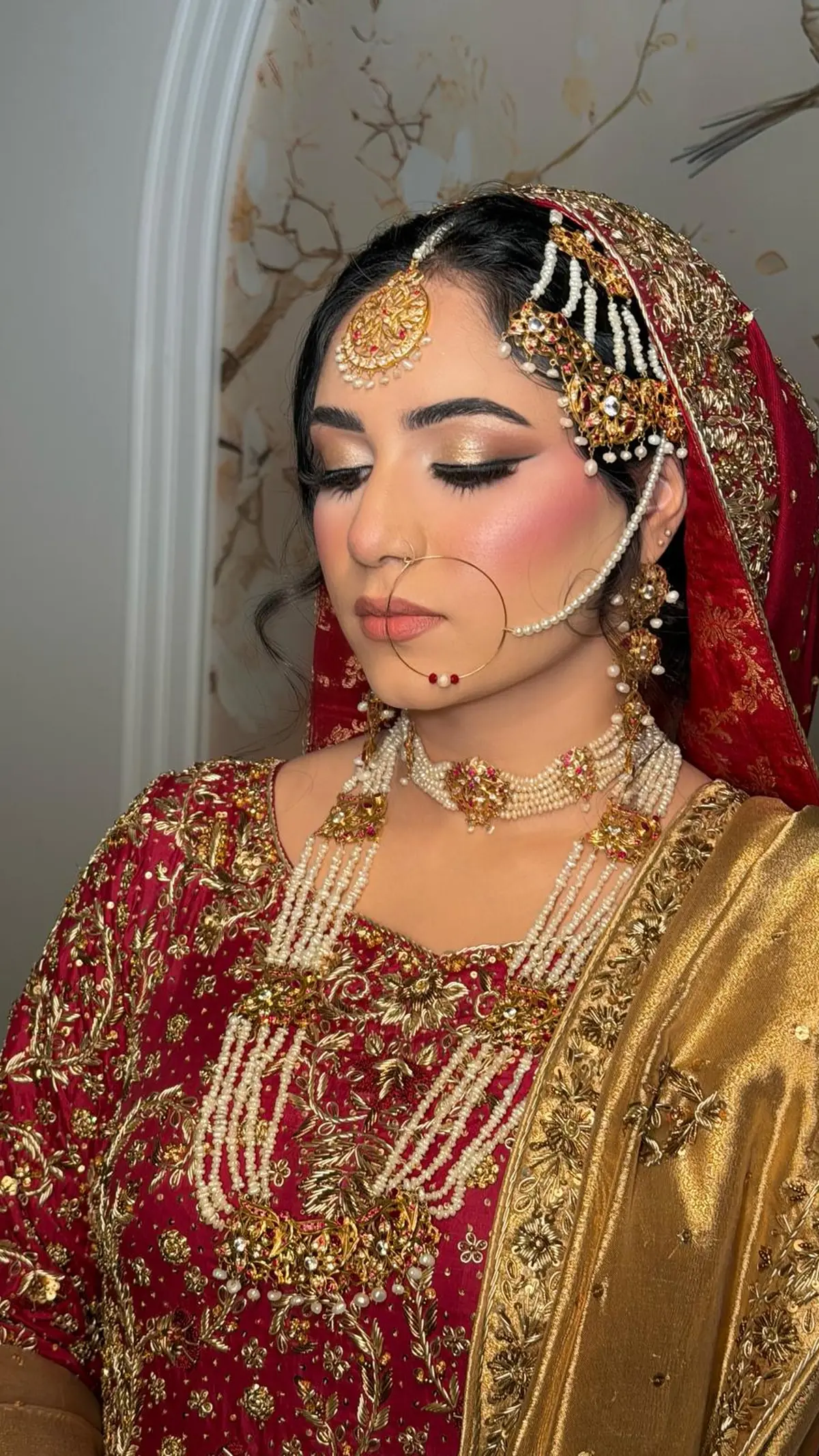 Bride in red and gold with a pearl choker and rani haar, a gold nath and jhoomar, and a gold tissue dupatta over one shoulder.