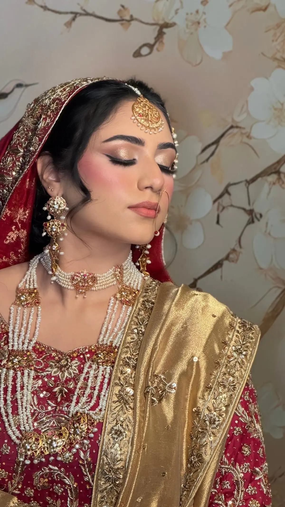Bride in red and gold with a pearl choker and layered rani haar, winged liner and a gold tissue dupatta, against a painted floral wall.