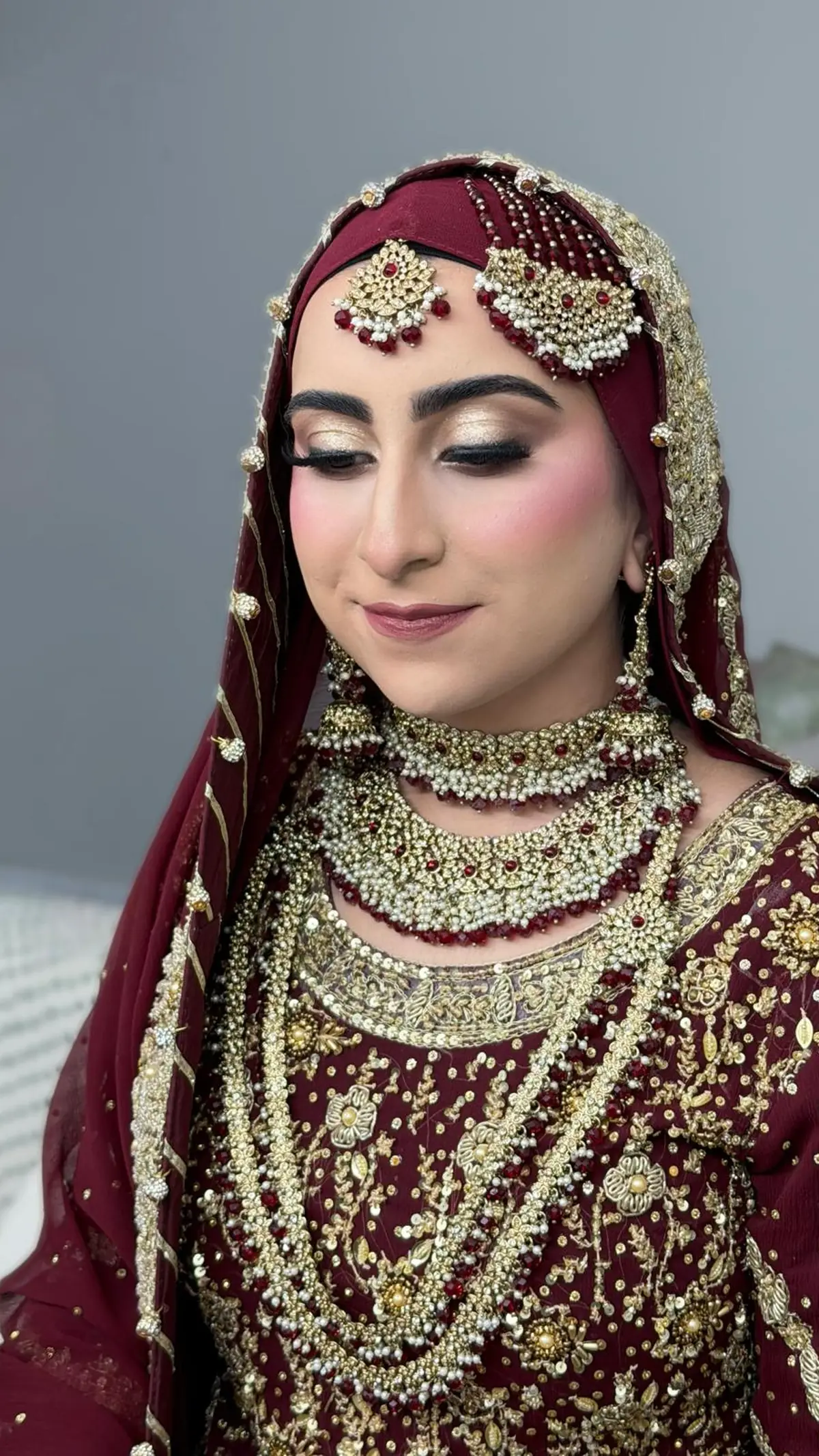 Hijabi bride in maroon and gold with a pearl jhoomar, layered pearl necklaces and a gold embellished dupatta.
