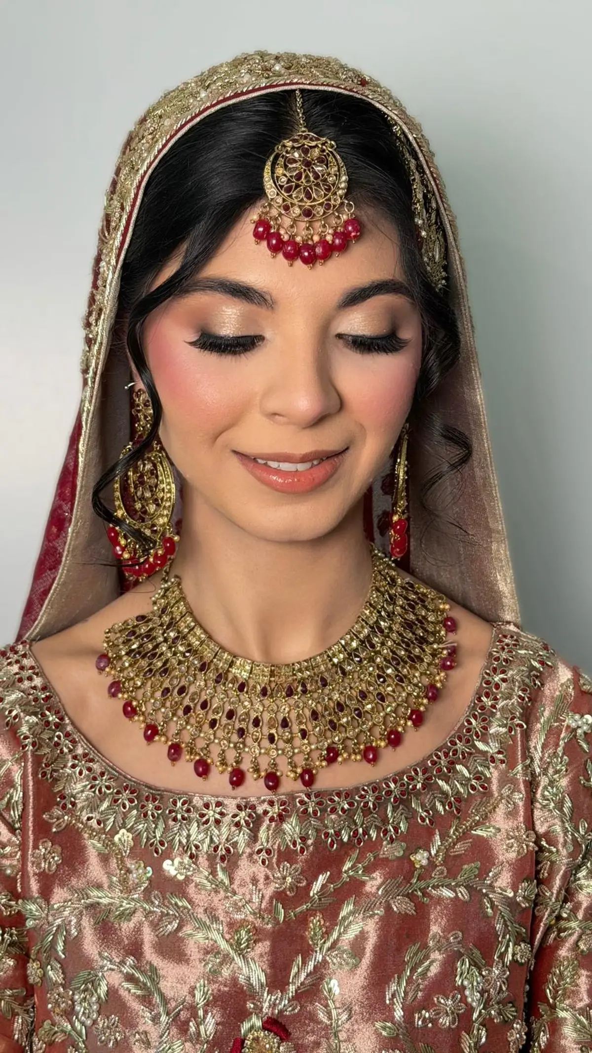Bride in rust and gold tilla work with a ruby drop necklace and maang tikka, soft curls under a red dupatta.