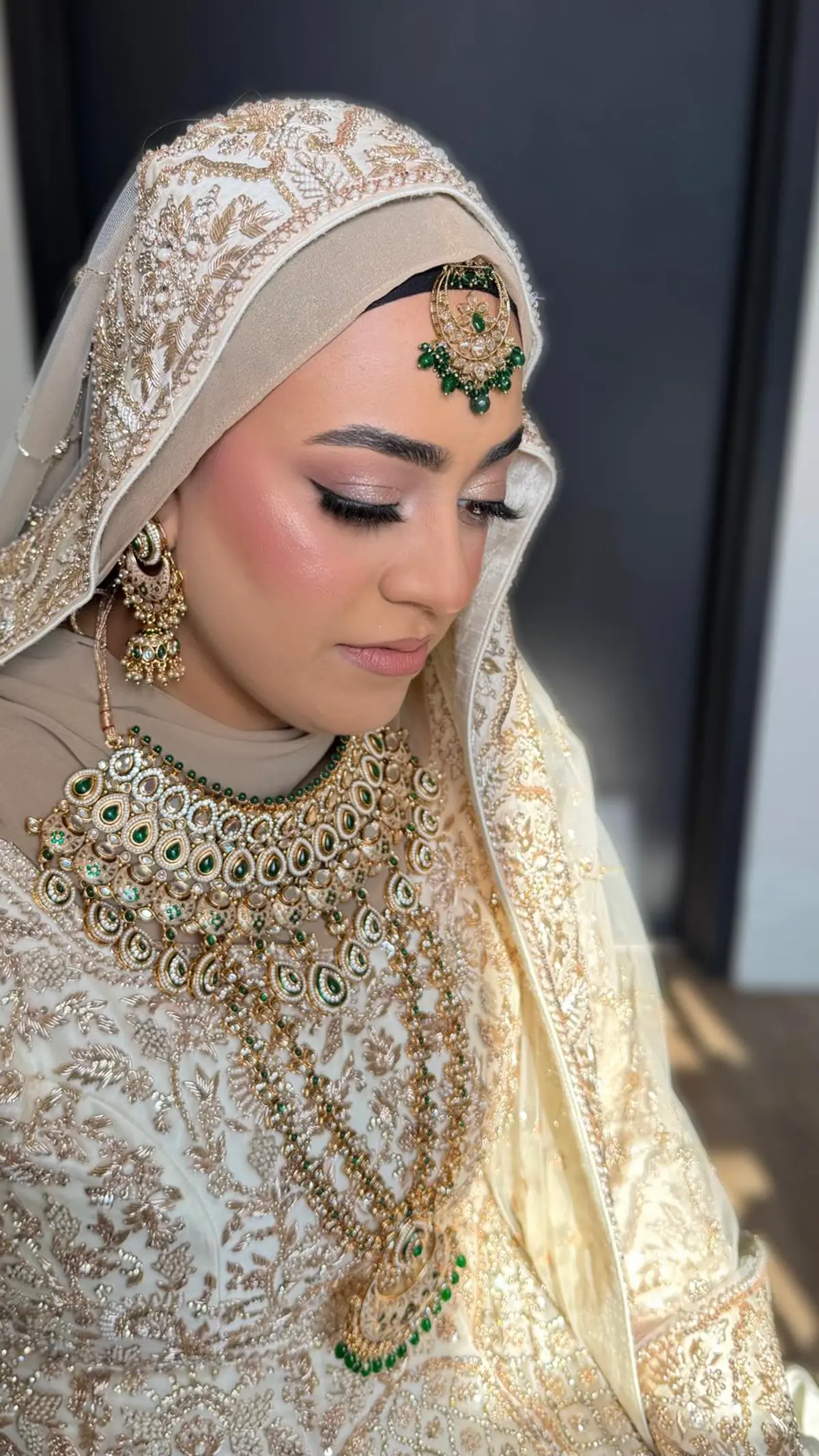 Hijabi bride in ivory and gold with an emerald kundan necklace and tikka, winged liner and a glowing finish.