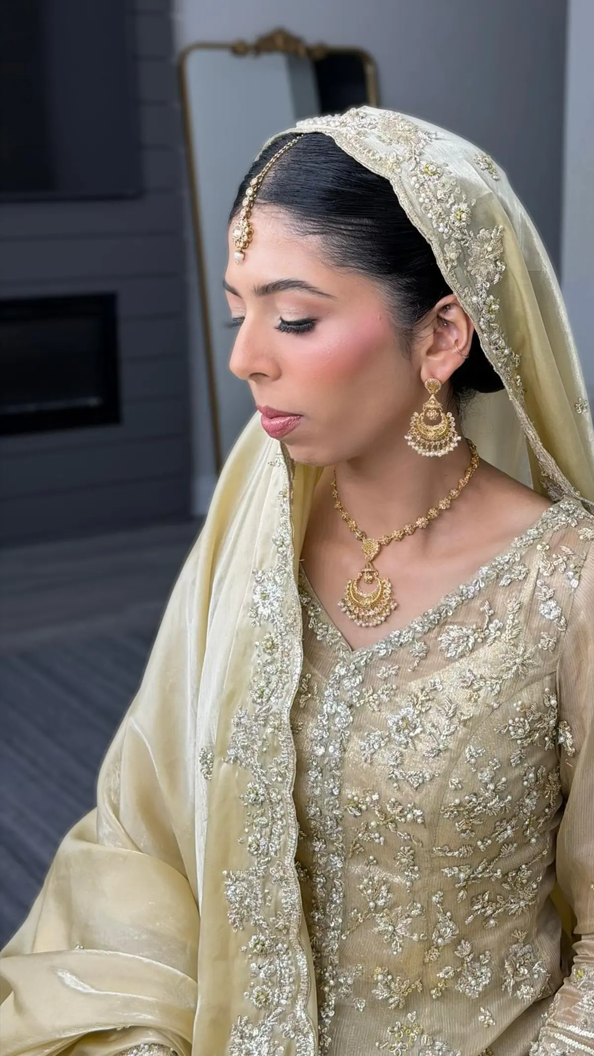 Bride in pale gold, seen in profile, with chandbali earrings, a slim tikka and a sleek low bun under her dupatta.